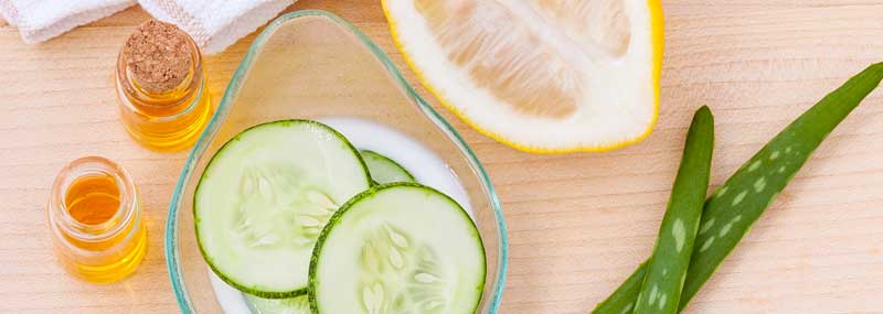 cucumber slices in a bowl, lemon, oils and aloe vera layed out on a light wooden table