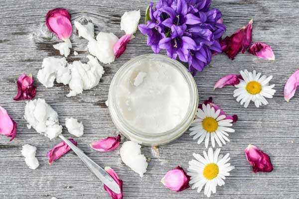 teaspoon full of coconut oil by a jar of coconut oil next to flowers and petals layed out on a grey wooden table