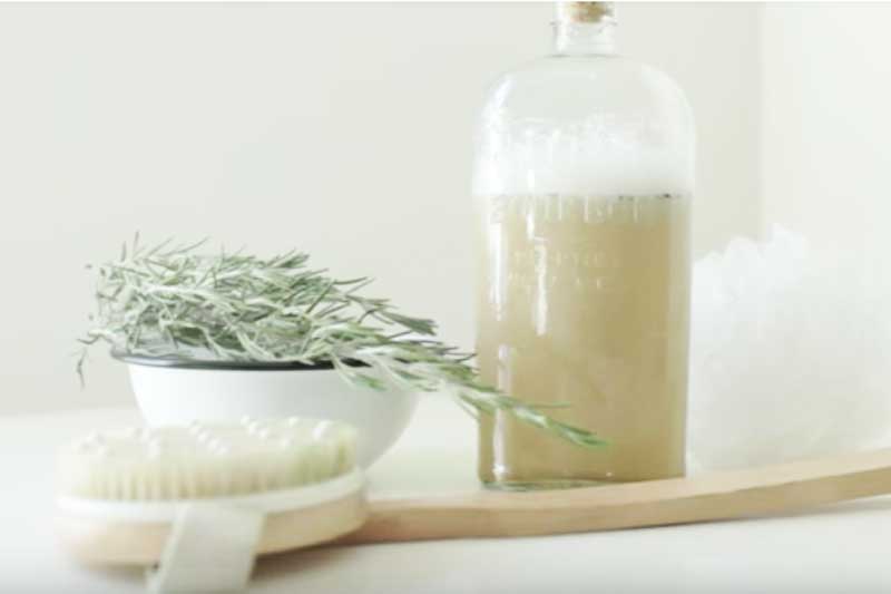 body wash in glass jug next to fresh rosemary and a body brush